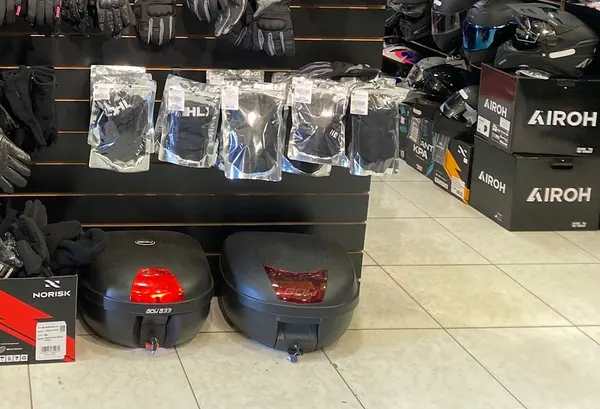 Dois baús de moto no chão da loja, ao lado de caixas de capacetes